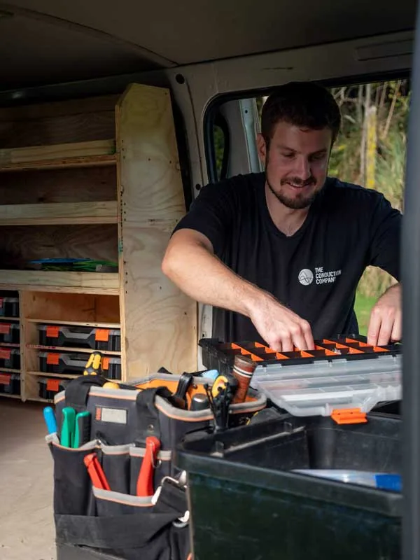 An electrician in a branded shirt selecting parts from the fitted shelving inside a work van