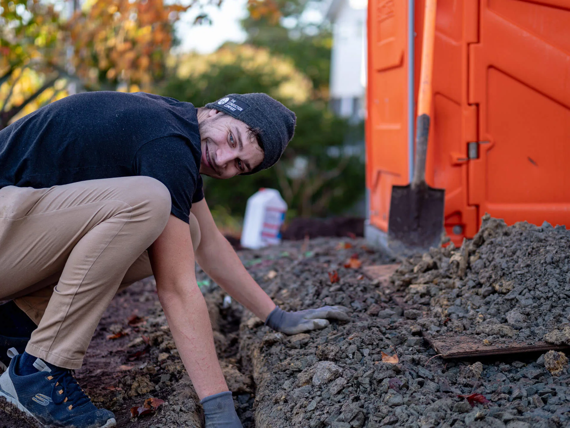 An electrician in a beanie kneeling to level gravel by hand beside an orange skip bin