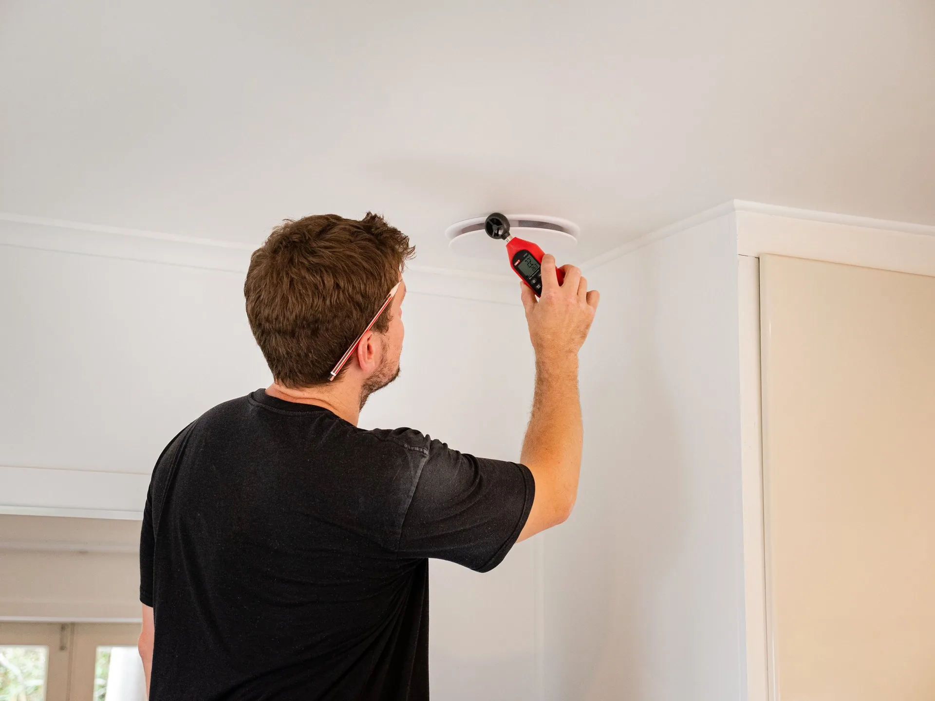An electrician reaching up to work on a ceiling fitting in a plain, lived in room