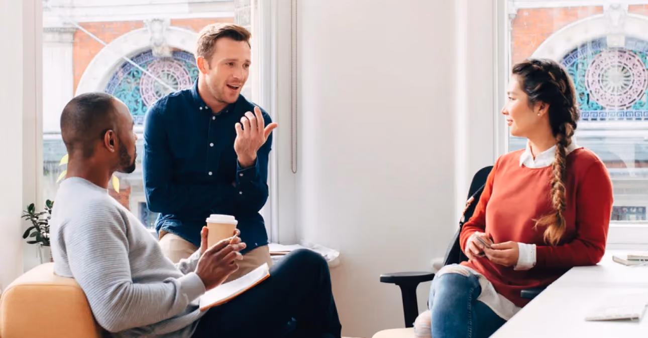 Three people sitting indoors having a discussion, one man gestures while the other man holds a coffee cup and notebook, and a woman listens attentively across from them.