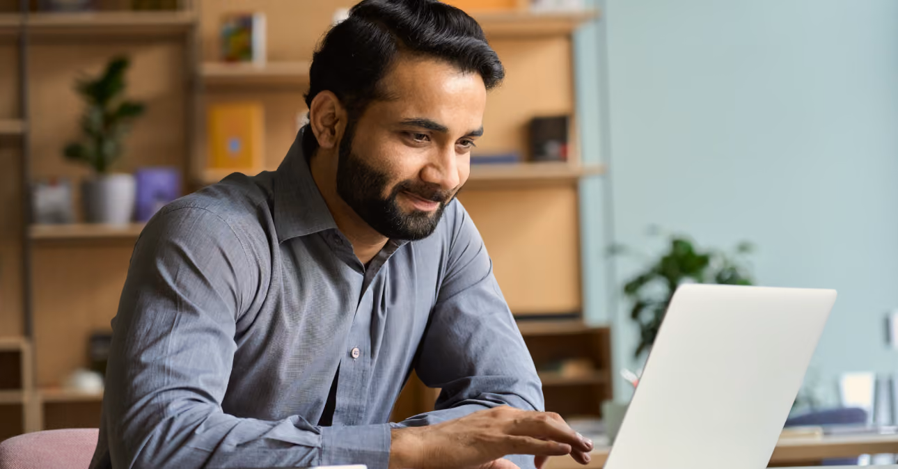 Man with dark hair and beard wearing a gray shirt, smiling and gesturing towards a laptop screen in a cozy room with shelves and plants in the background.