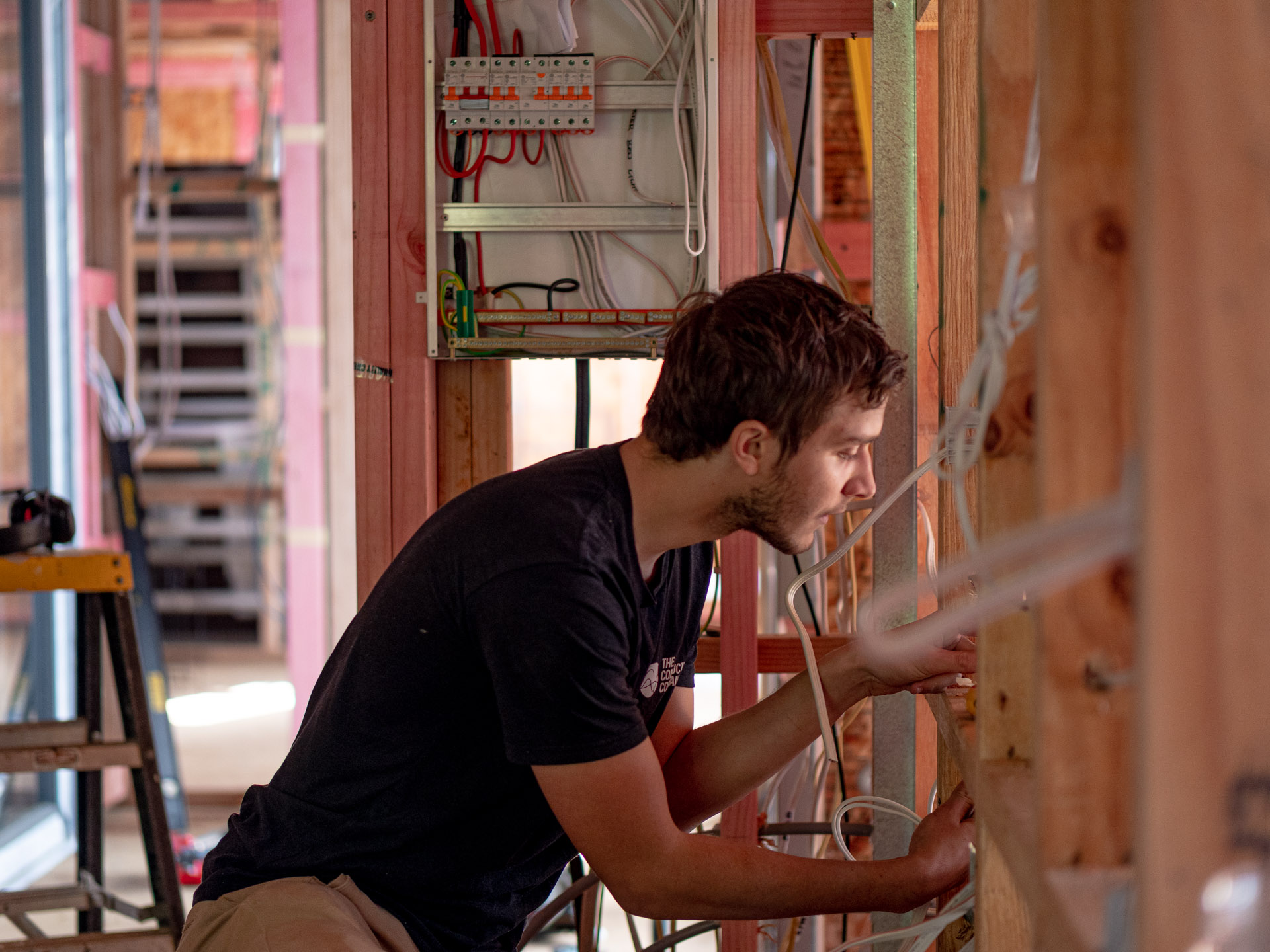 An electrician working on a switchboard set into an unlined timber-framed wall