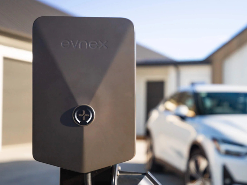 A dark post-mounted EV charger with a white car and garage behind it