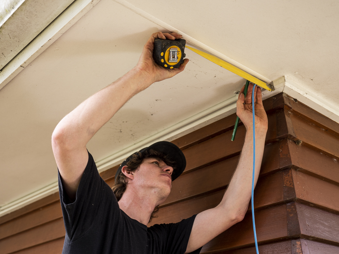 An electrician measuring along a soffit with a tape measure, blue cable in hand