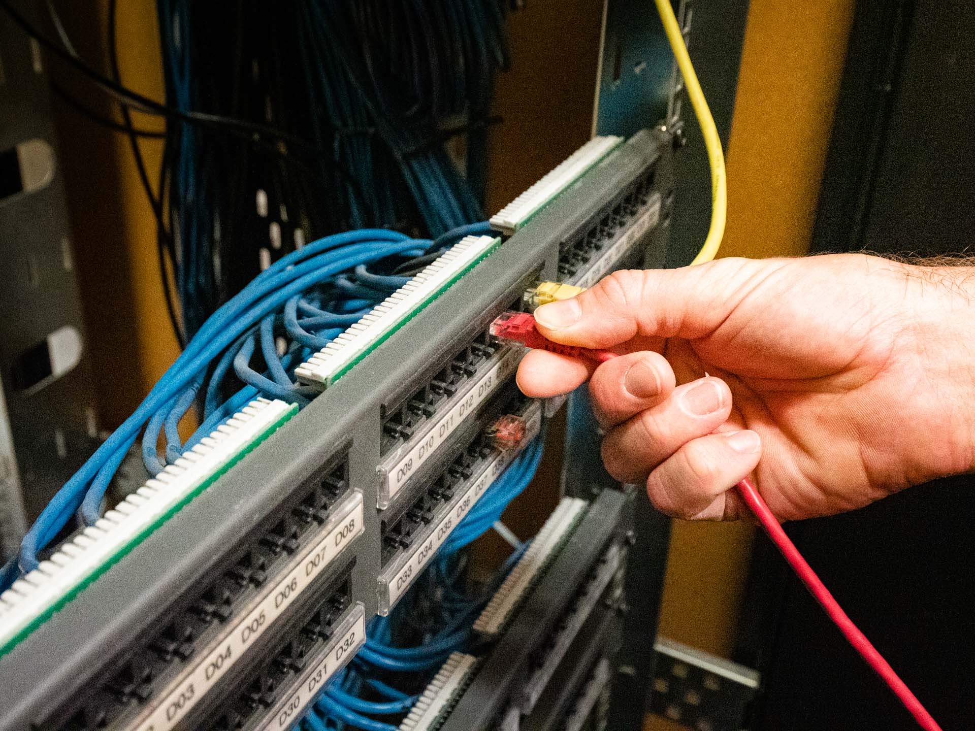 A hand plugging a red patch lead into a patch panel filled with blue network cables