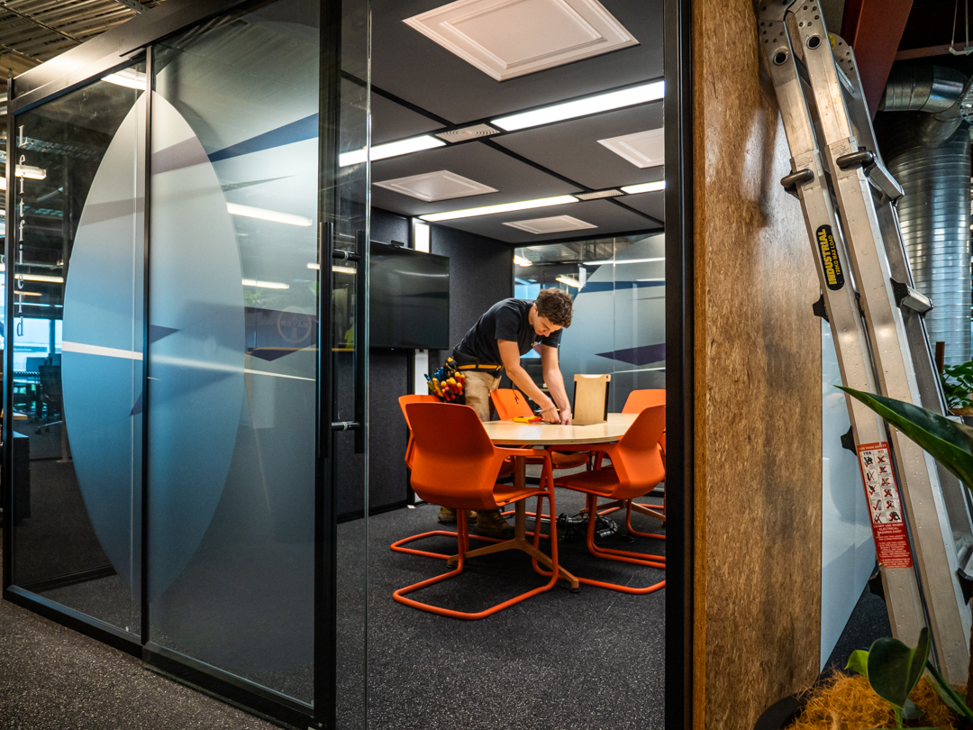 An office with glass partitions and orange chairs, an electrician working at a table