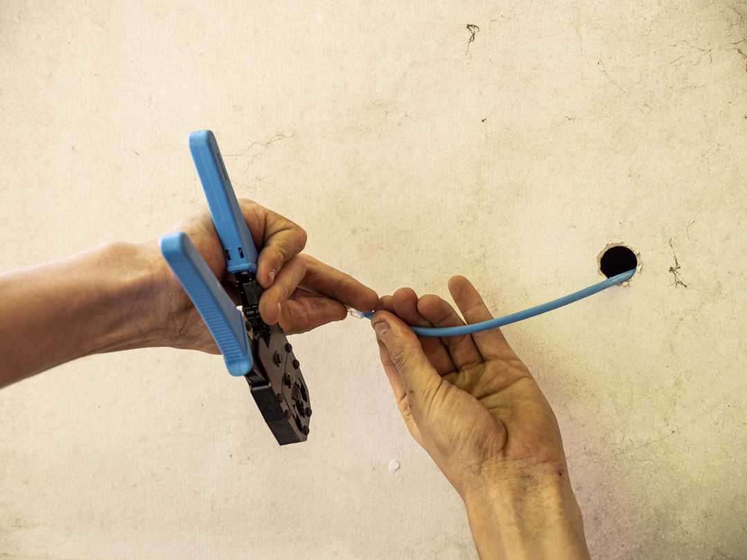 Hands crimping a blue network cable beside an opening cut in a wall