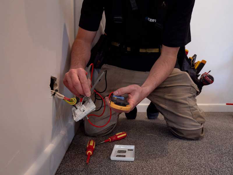 An electrician kneeling to test a wall socket with a hand-held tester