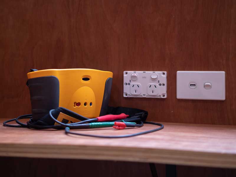 A yellow electrical tester on a bench beside two socket outlets and test leads