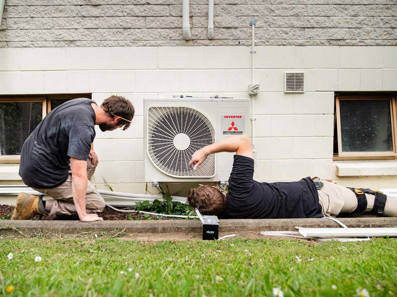 Two electricians fitting an outdoor heat pump unit against a block wall
