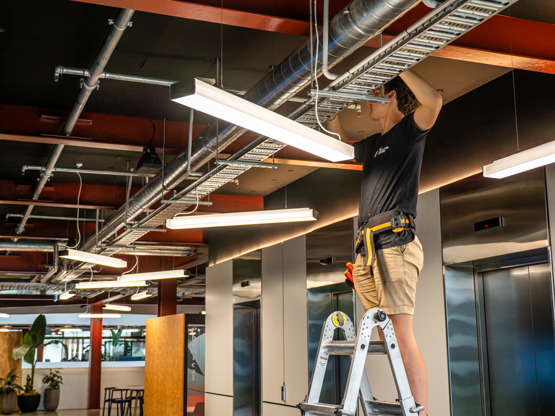 Electrician on a step ladder fitting a linear LED batten to an exposed services ceiling in an open plan office.