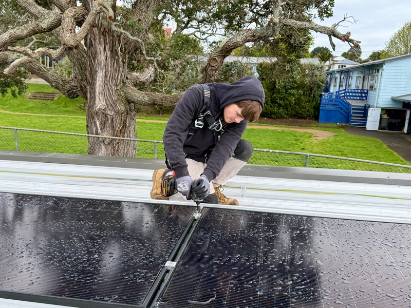 An electrician in a safety harness fastening a solar panel to roof rails with a cordless drill