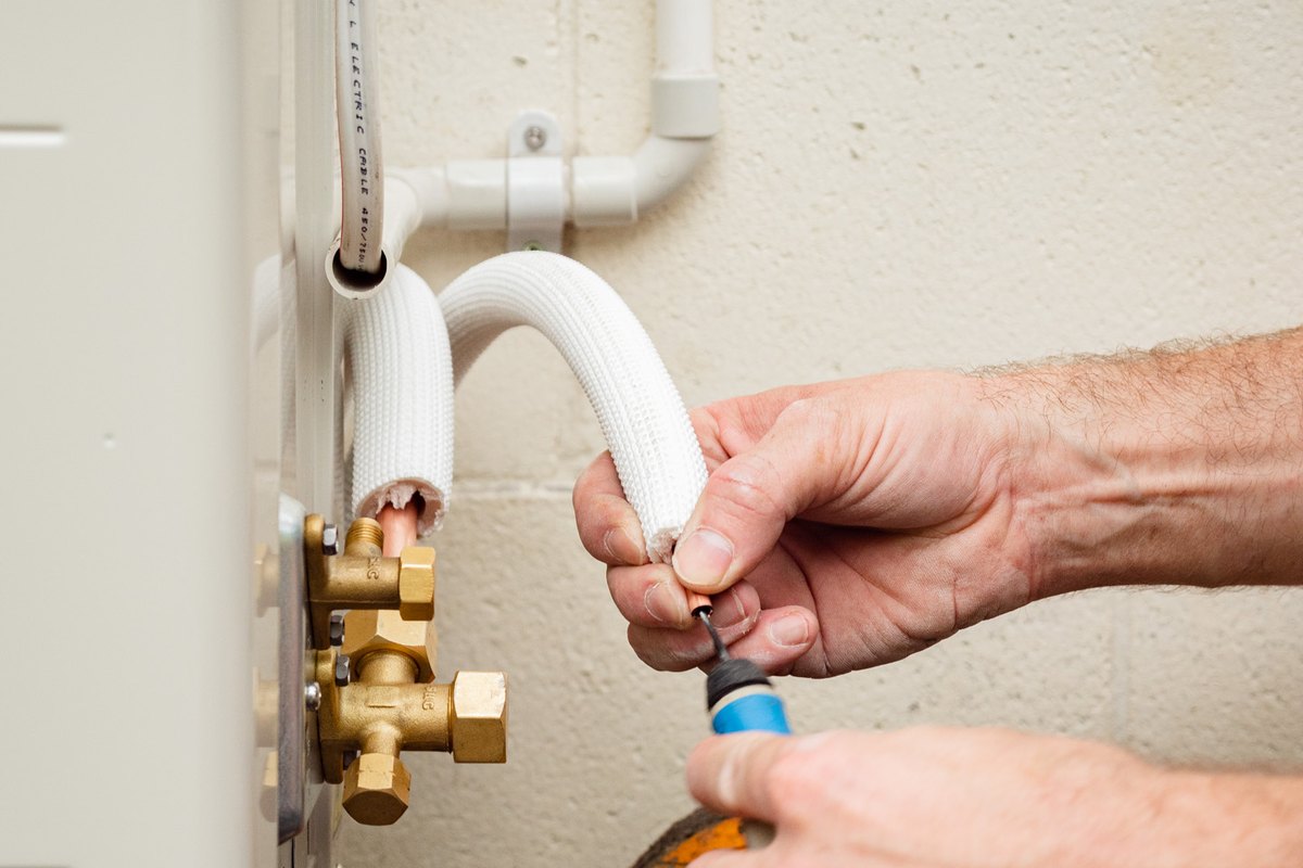 Two hands connecting the brass service valves at the base of an outdoor heat pump unit.