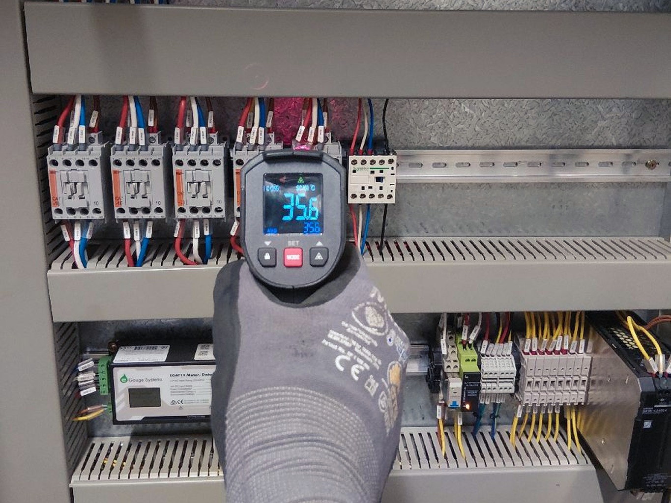 A gloved hand holding an infrared thermometer up to a row of contactors in a control panel, the screen showing the reading