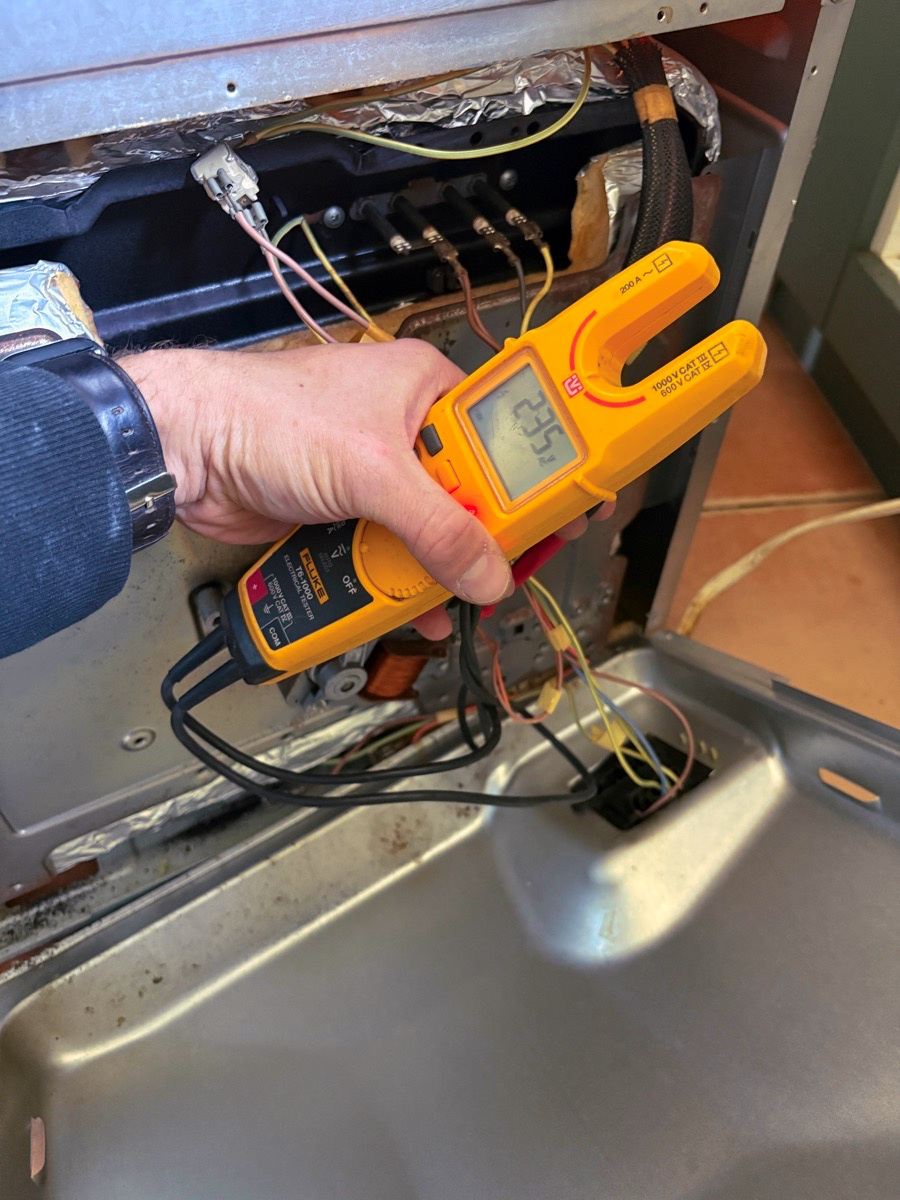 A hand holding an electrical tester at the opened back of an oven, the screen showing the voltage reading