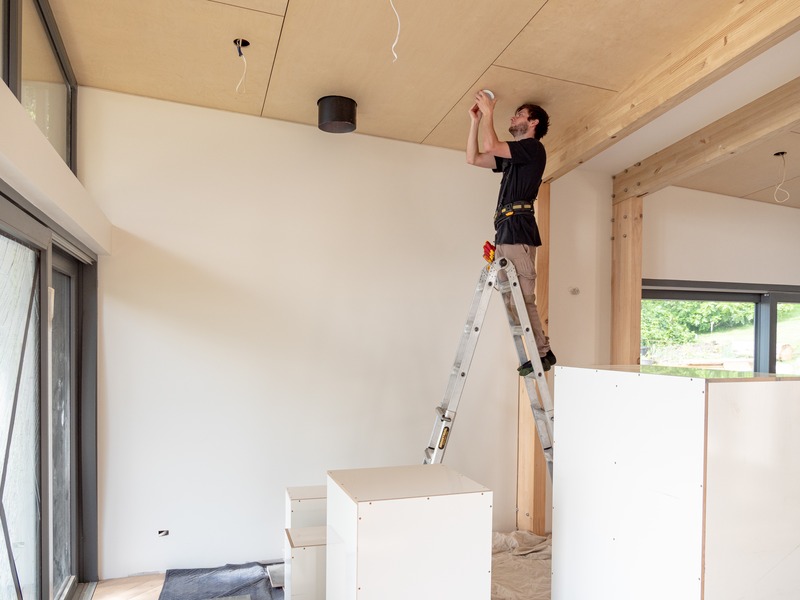 An electrician on a ladder fitting a downlight into the plywood ceiling of a newly built room
