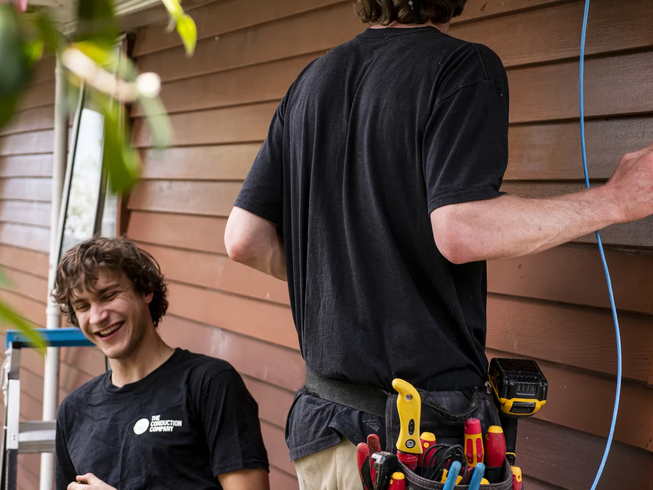 Two electricians at the weatherboard wall of a house, one with a loaded tool belt, blue cable running up to the eaves