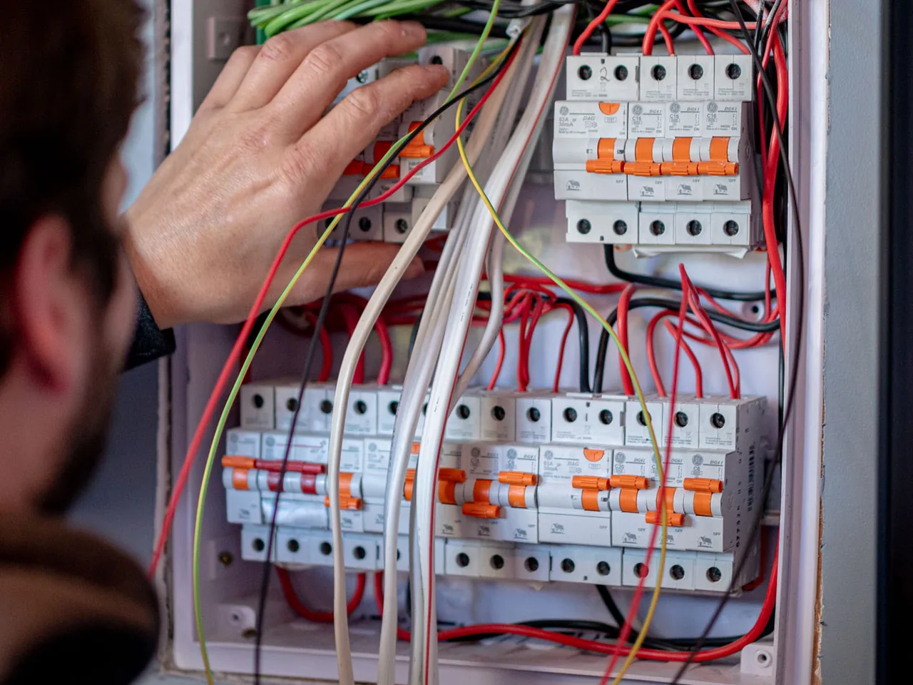 Electrician's hand on a row of circuit breakers inside an open switchboard full of red and white wiring