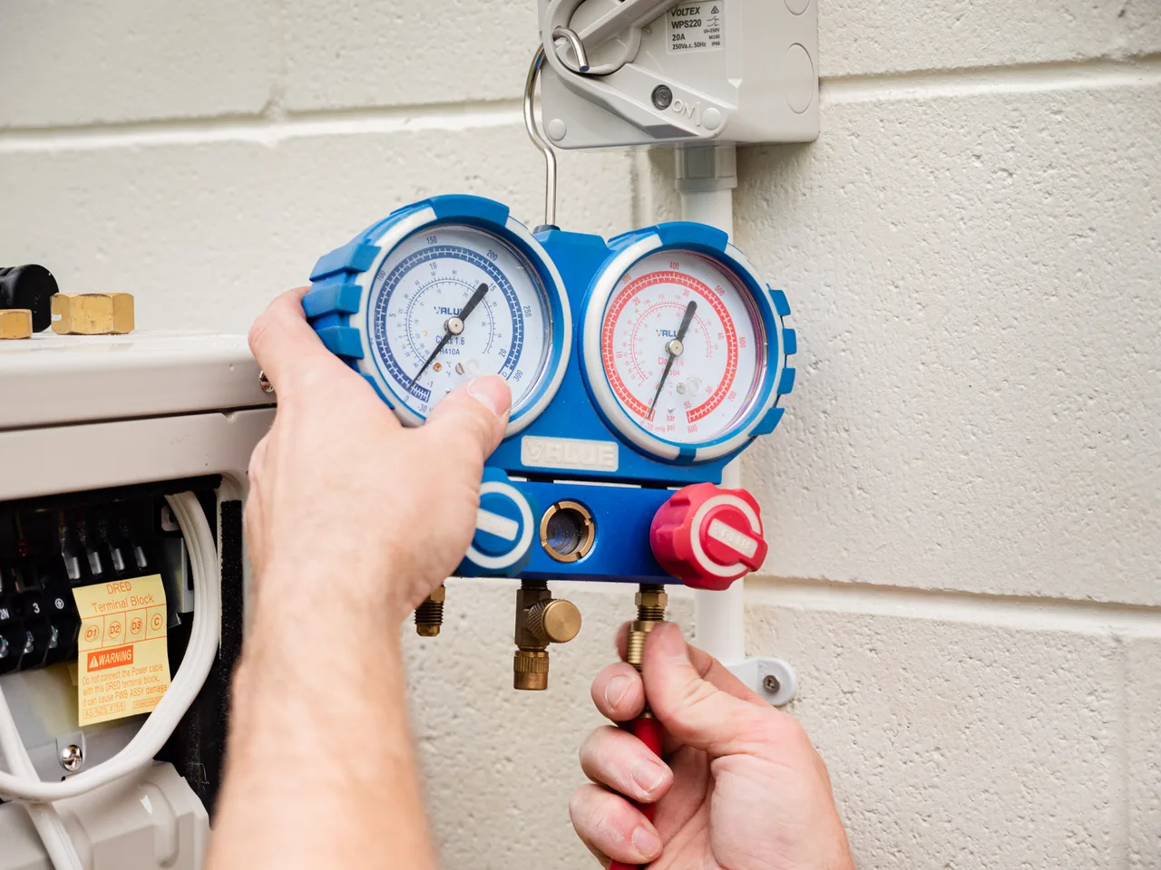 Hands holding a twin gauge refrigeration manifold beside the open terminal cover of an outdoor unit