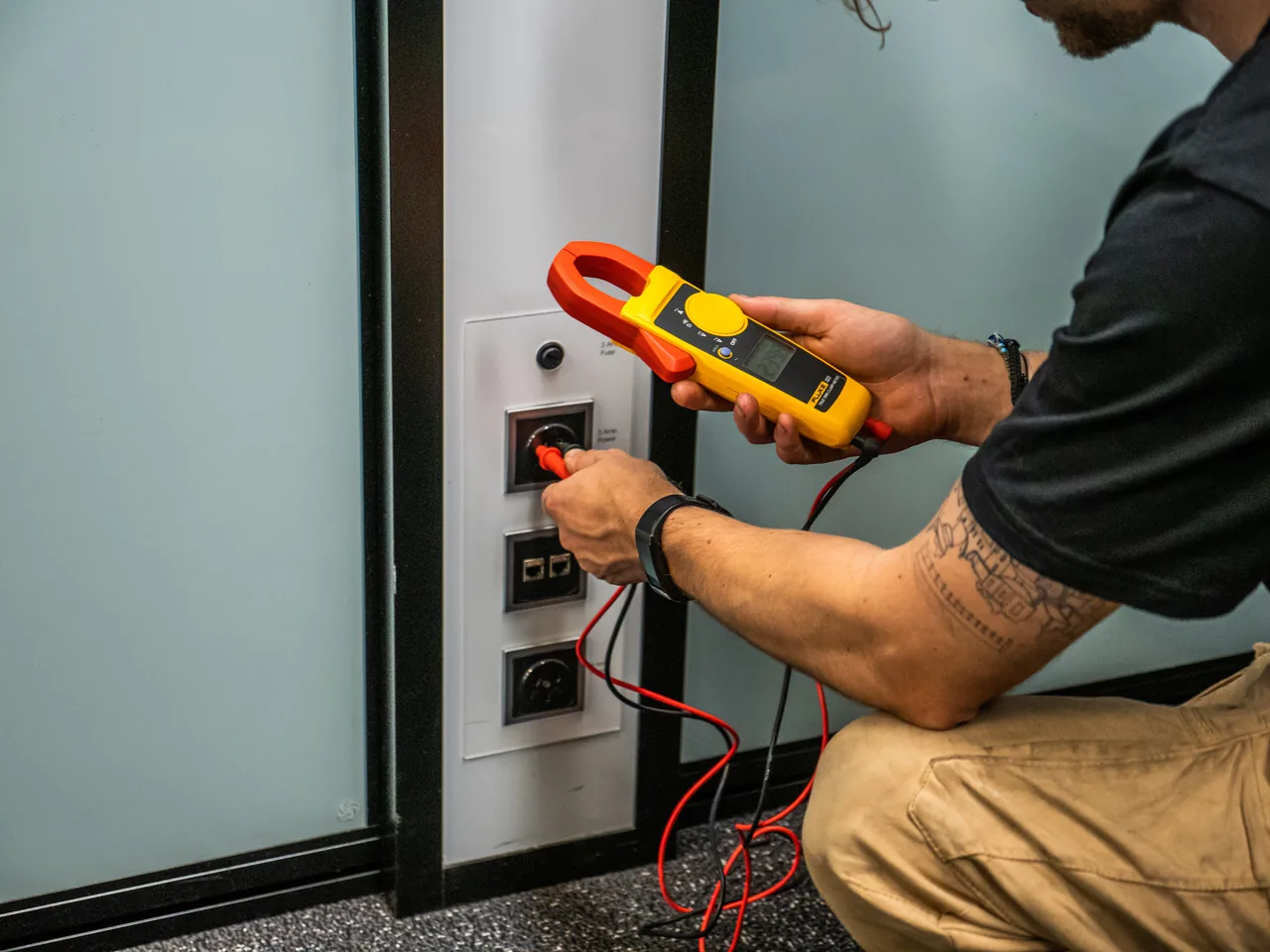 Hands testing a wall socket with a yellow clamp meter and red probes, a glass office partition behind