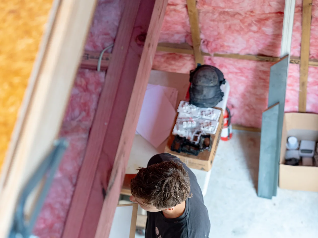 Looking down through open framing at an electrician among boxes of fittings, pink insulation batts in the walls