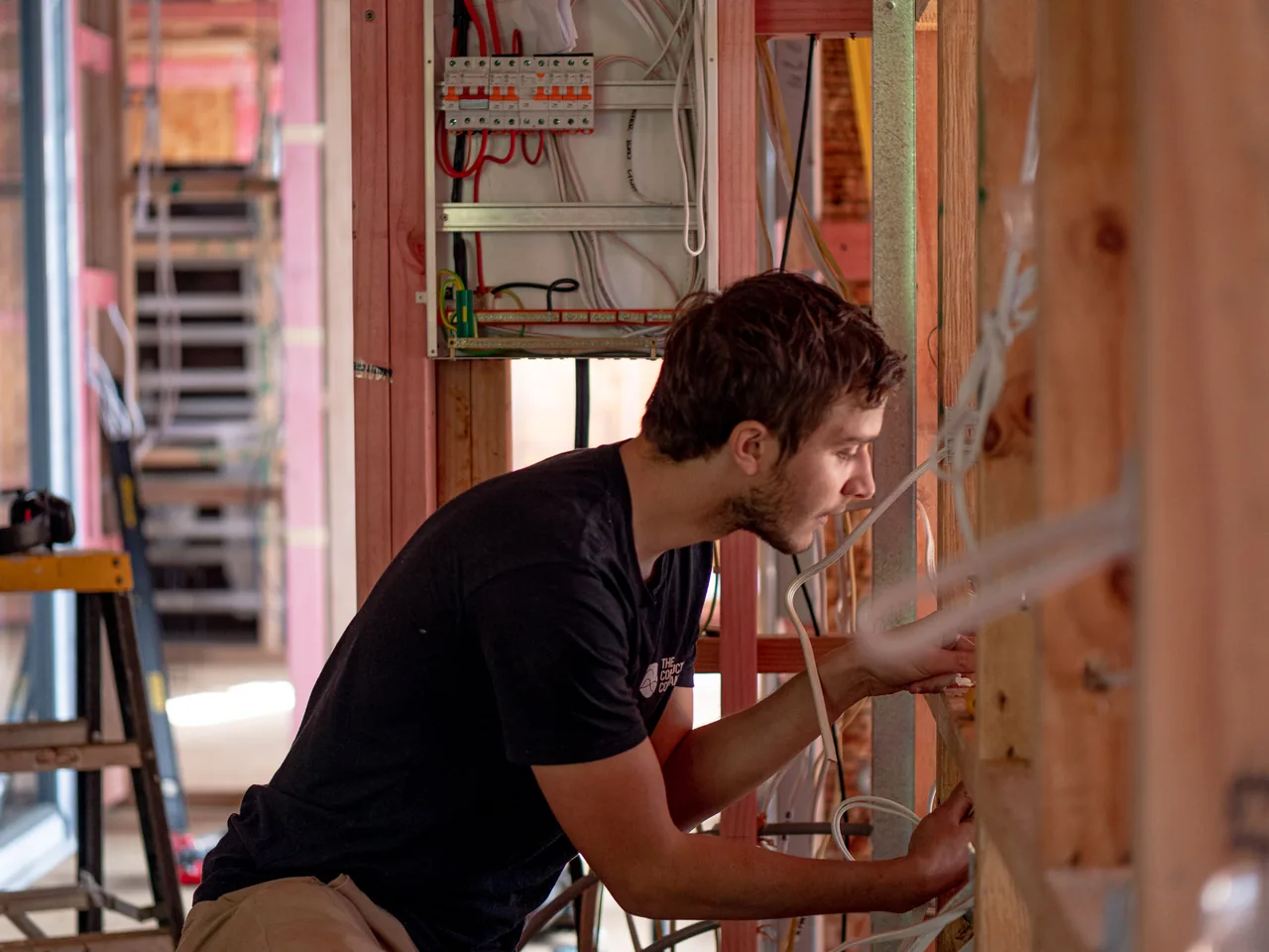 Electrician working cables through open timber framing, with a new switchboard set into the wall frame behind him