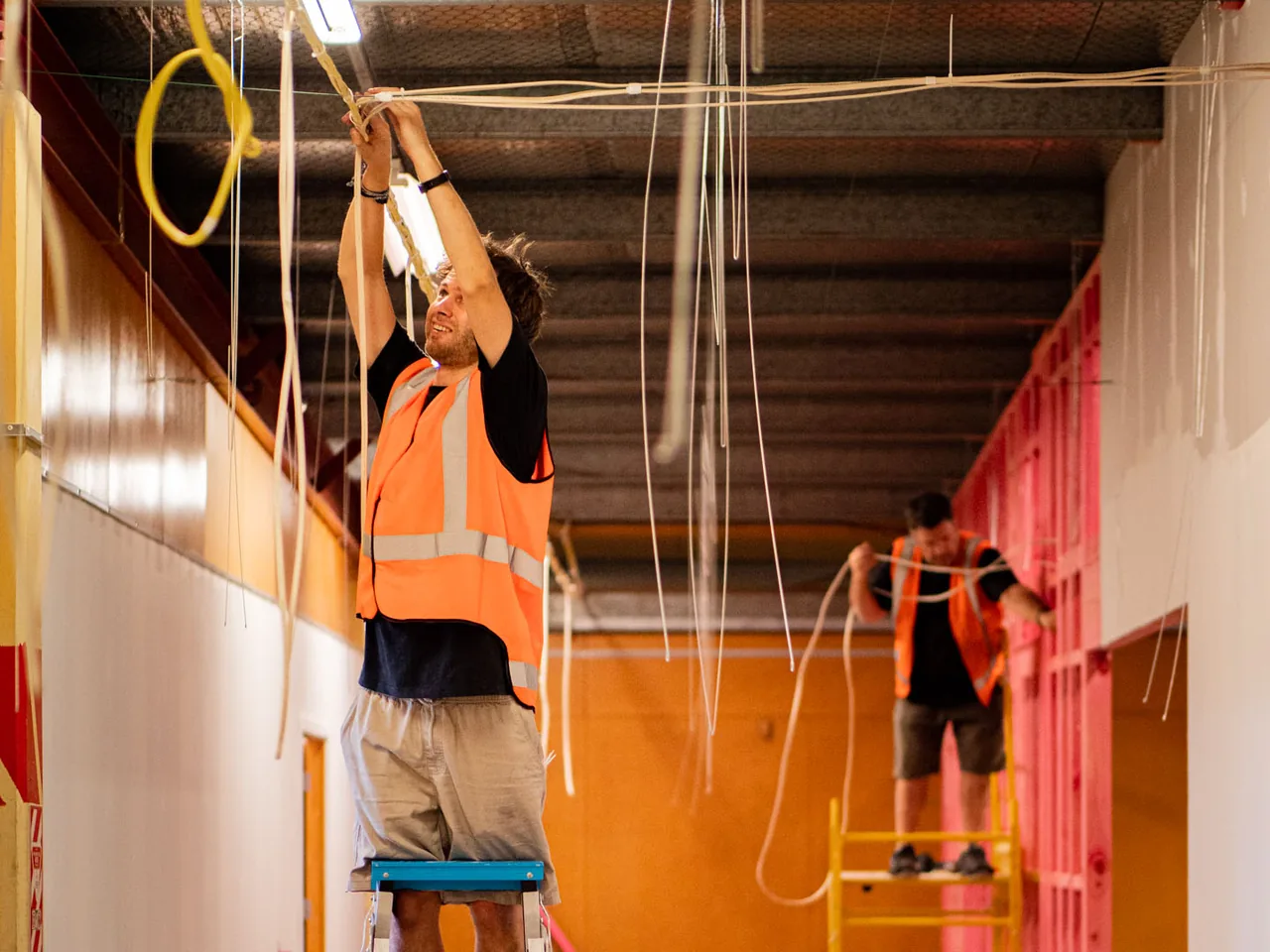 Electrician on a step ladder pulling cable in a commercial fit-out corridor, cables hanging from the exposed ceiling
