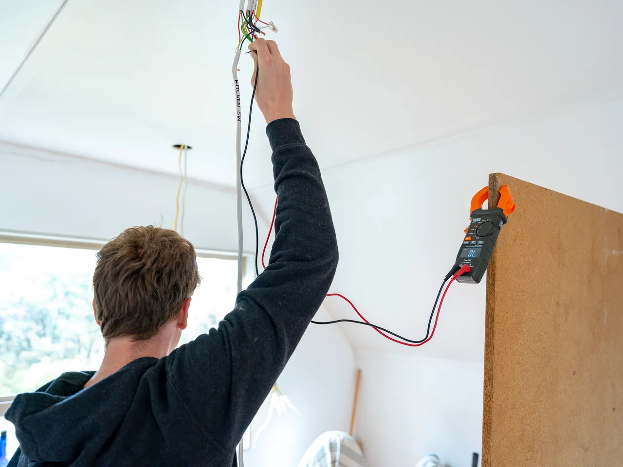 Electrician reaching up to a ceiling cable with test leads running to a clamp meter hung on a board