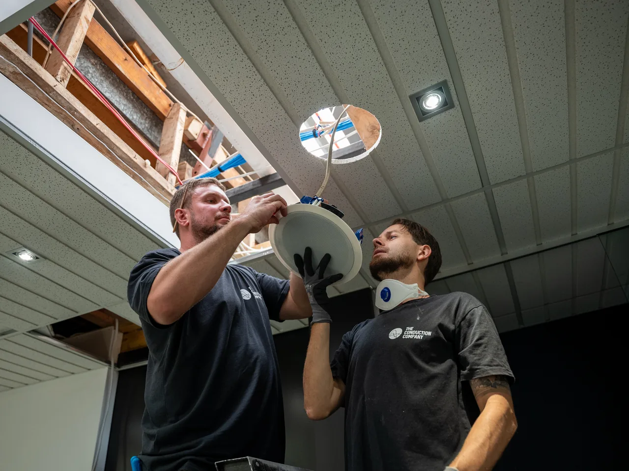 Two electricians lifting a round in-ceiling speaker up into a hole cut in a ceiling tile