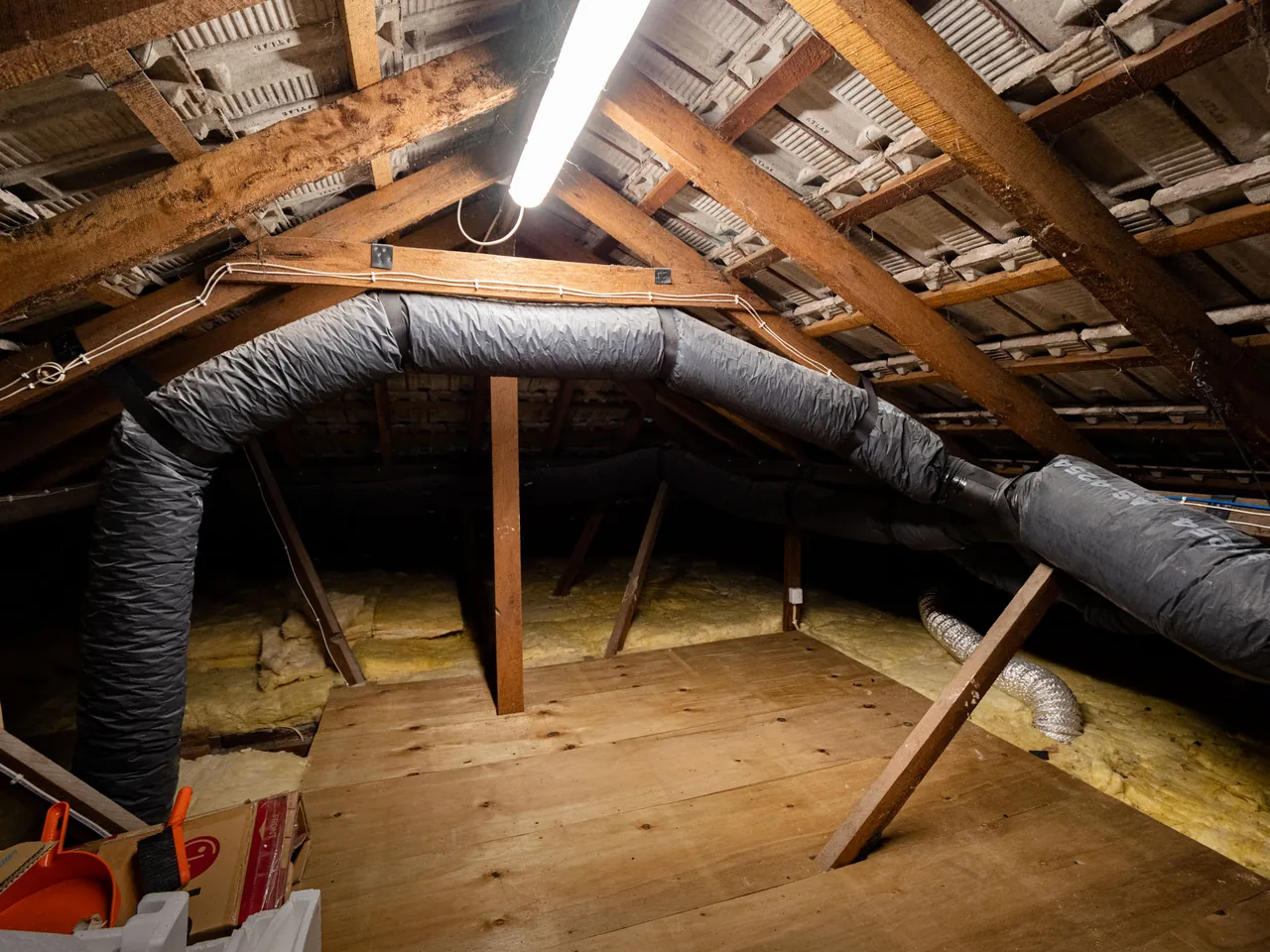 Insulated flexible ducting running across the ceiling joists of a roof space, lit by a work light
