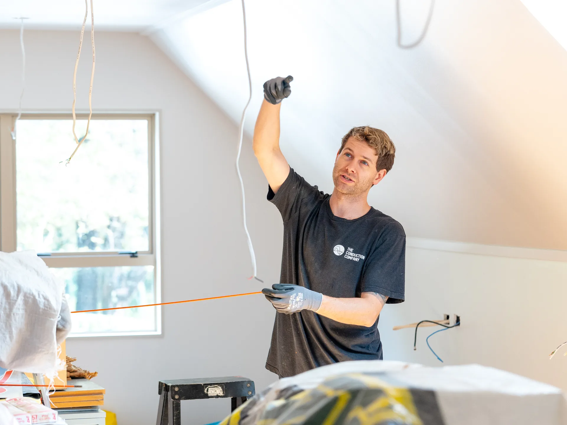 Electrician in a lived-in attic reaching up to a ceiling point while running new cable with a rod, furniture under sheets