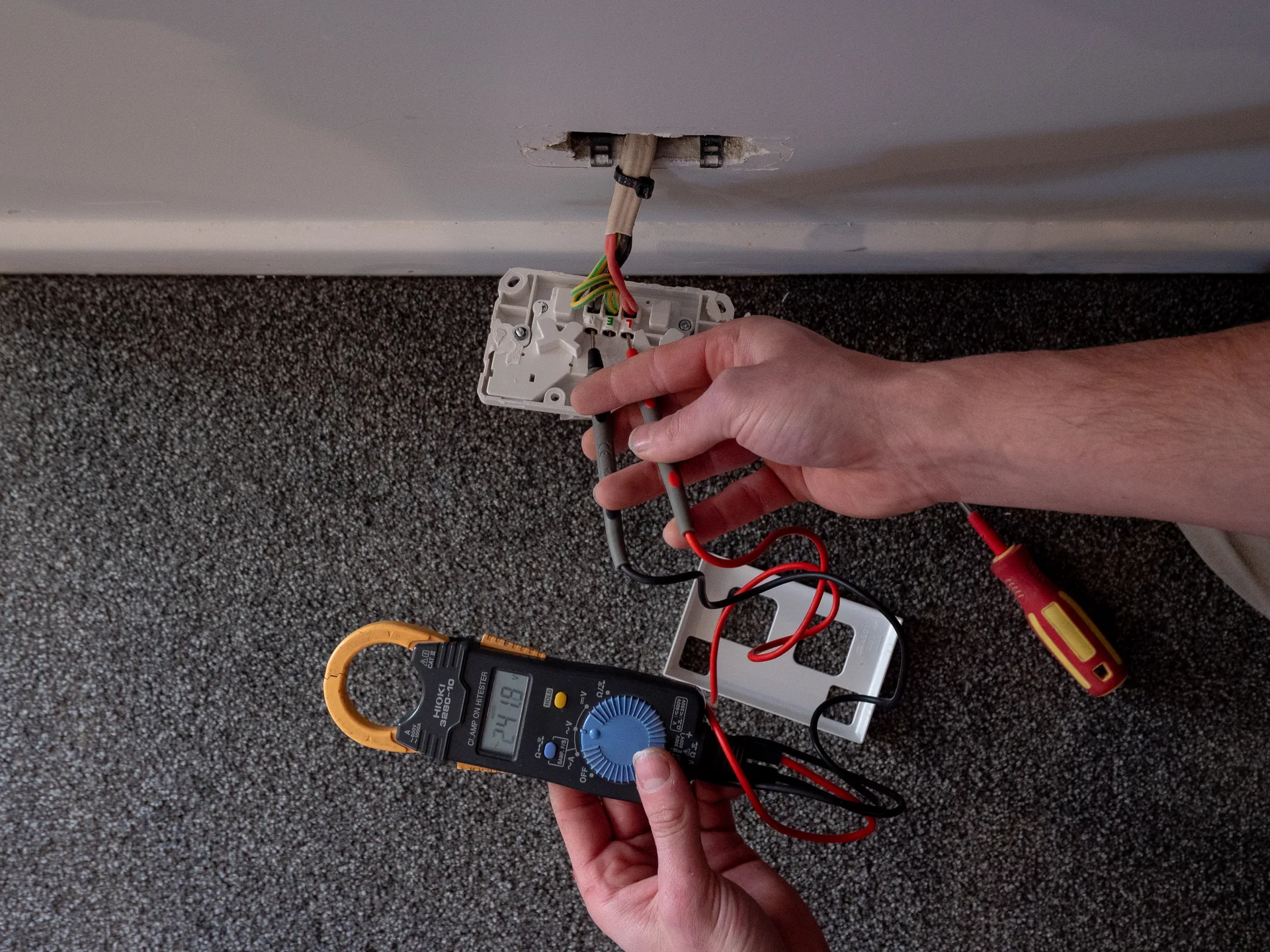 Hands testing a power point pulled off the wall above the skirting, with a clamp meter reading mains voltage on the carpet