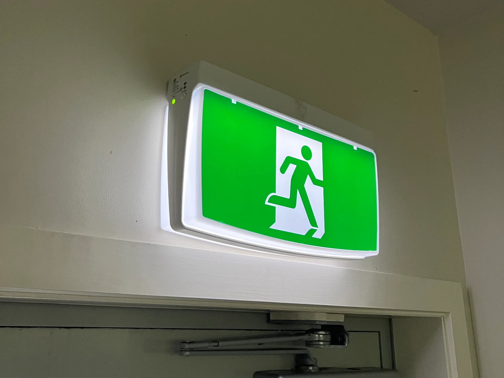 Lit green running man exit sign mounted on the wall above a door with a door closer