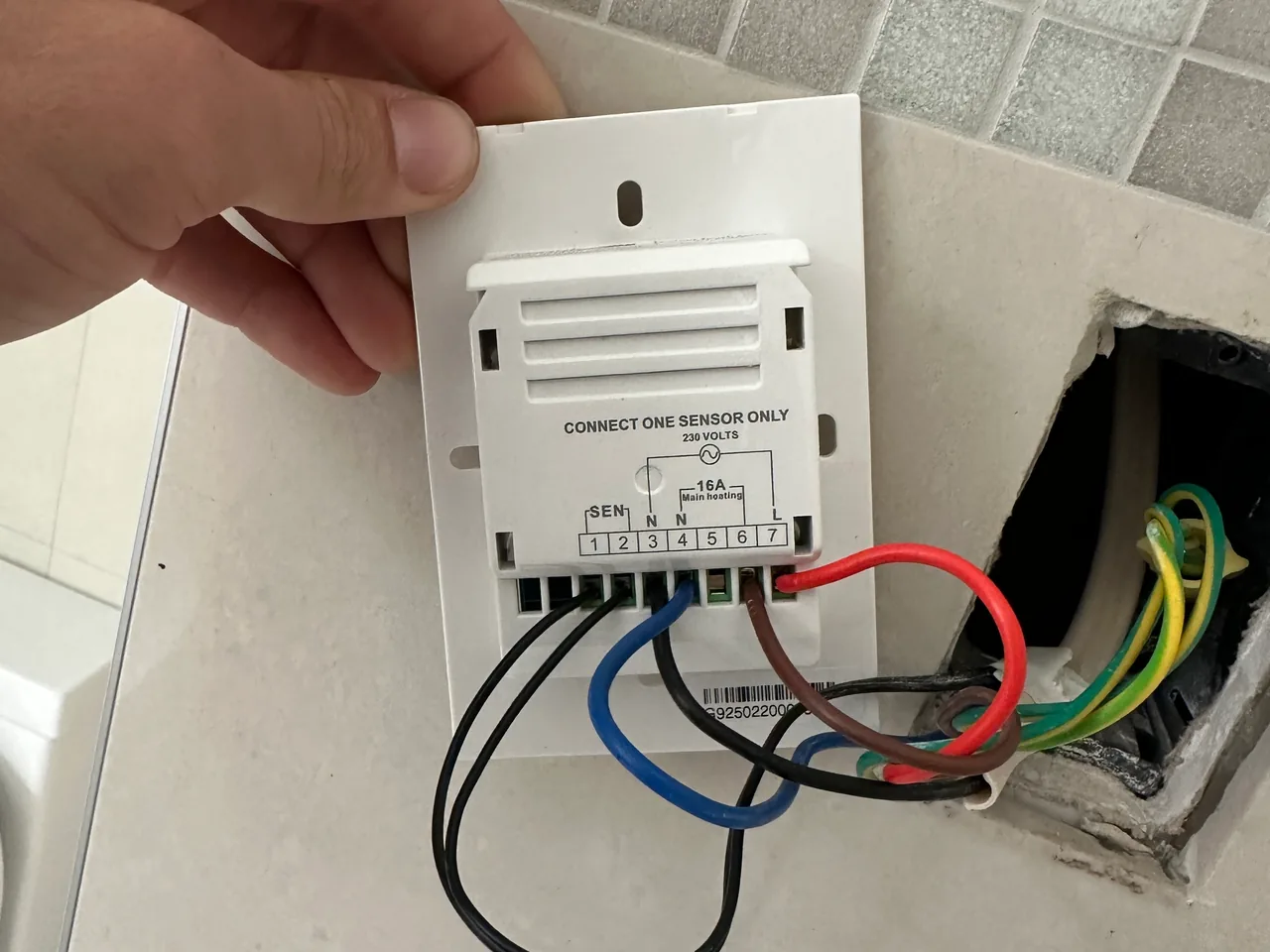 Hand holding the back of a floor heating thermostat as its wires are connected into a wall box in bathroom tiles