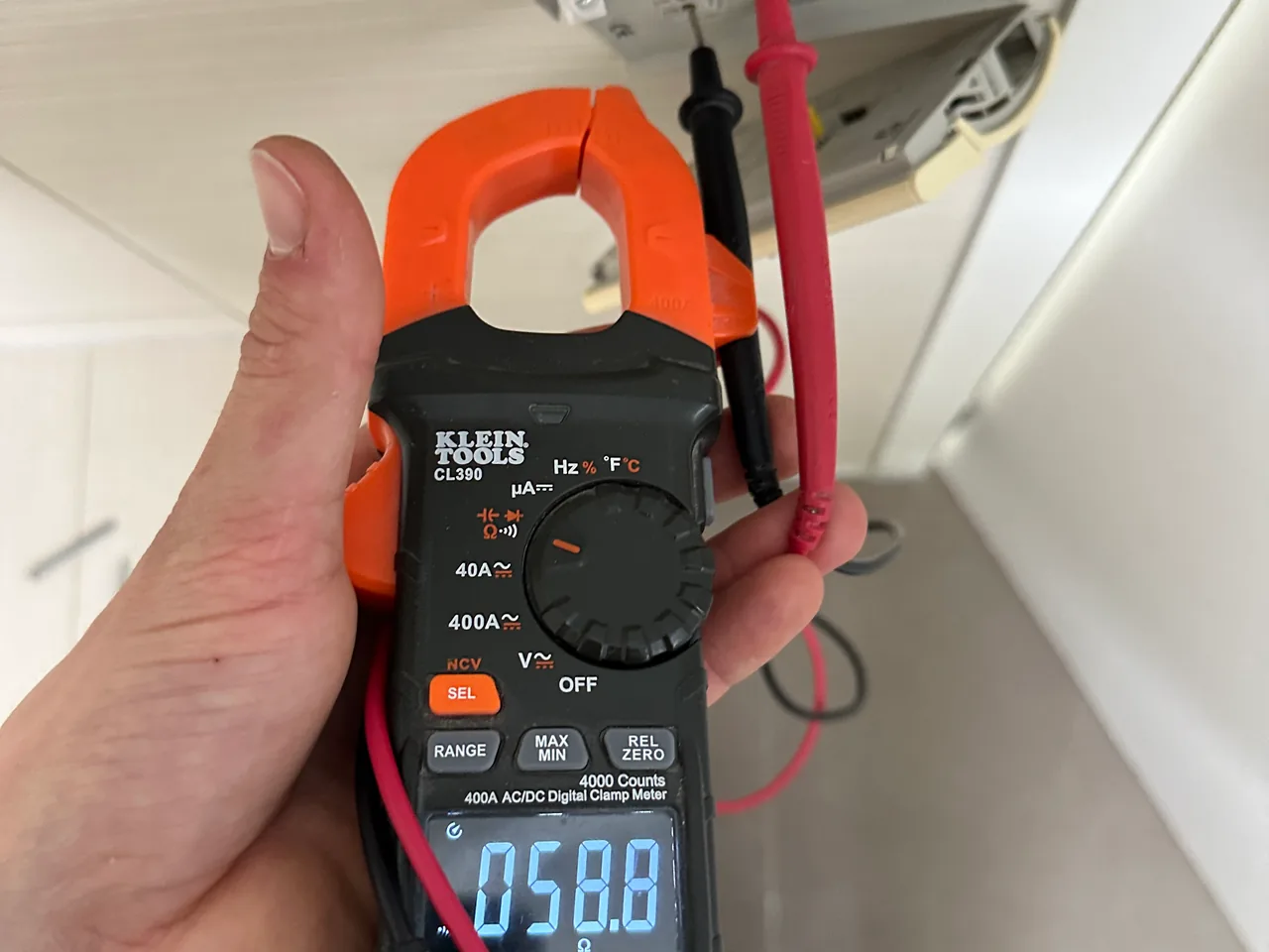 Hand holding a clamp meter reading 58.8 ohms, its test leads running to the wires of an opened floor heating thermostat