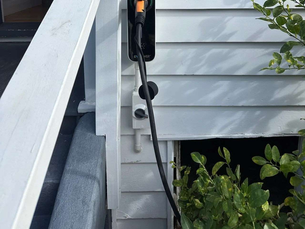 A black EV charger unit mounted on white weatherboard cladding beside a porch, its cable coiled below.