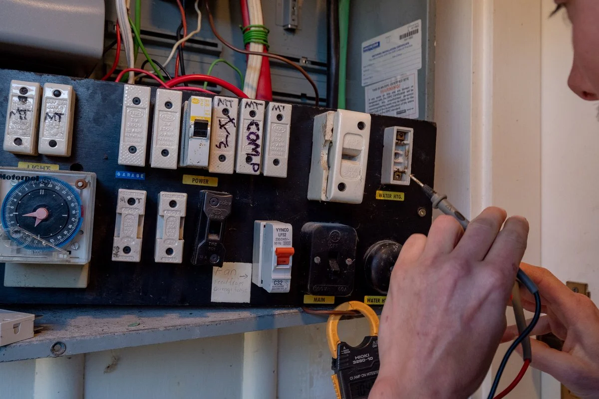 A hand with a test probe at an old switchboard: ceramic fuses, a mechanical time clock and mismatched breakers.