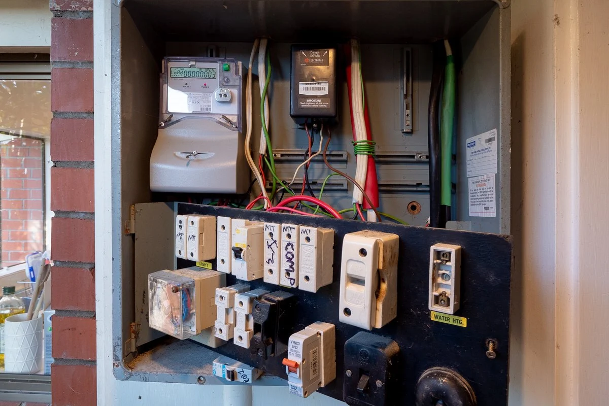 An old meter box recessed in a wall, with a time clock, old fuses and a loose hanging cable.