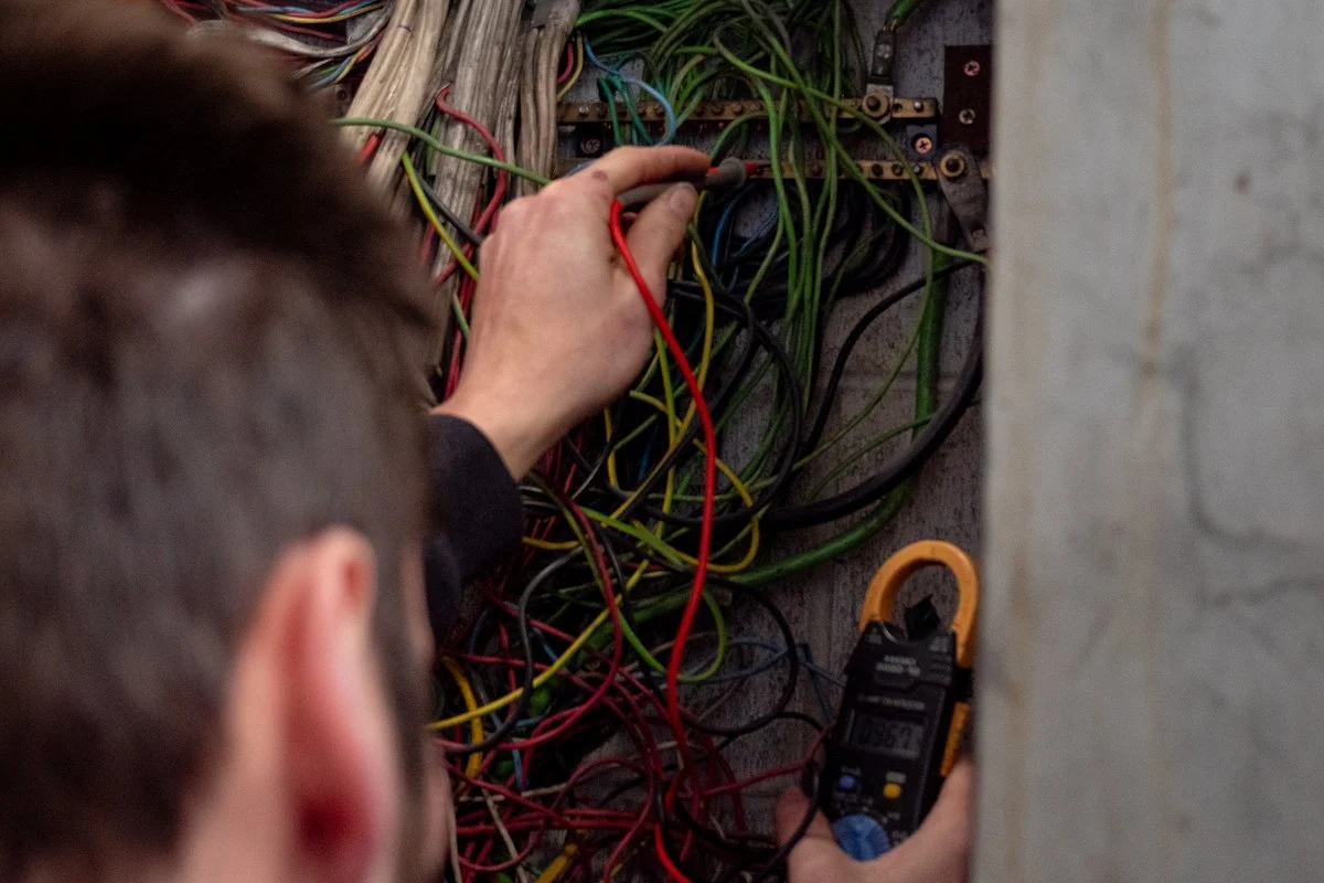 A test probe in a dense mass of old wiring at a distribution board, a clamp meter hanging beside it.