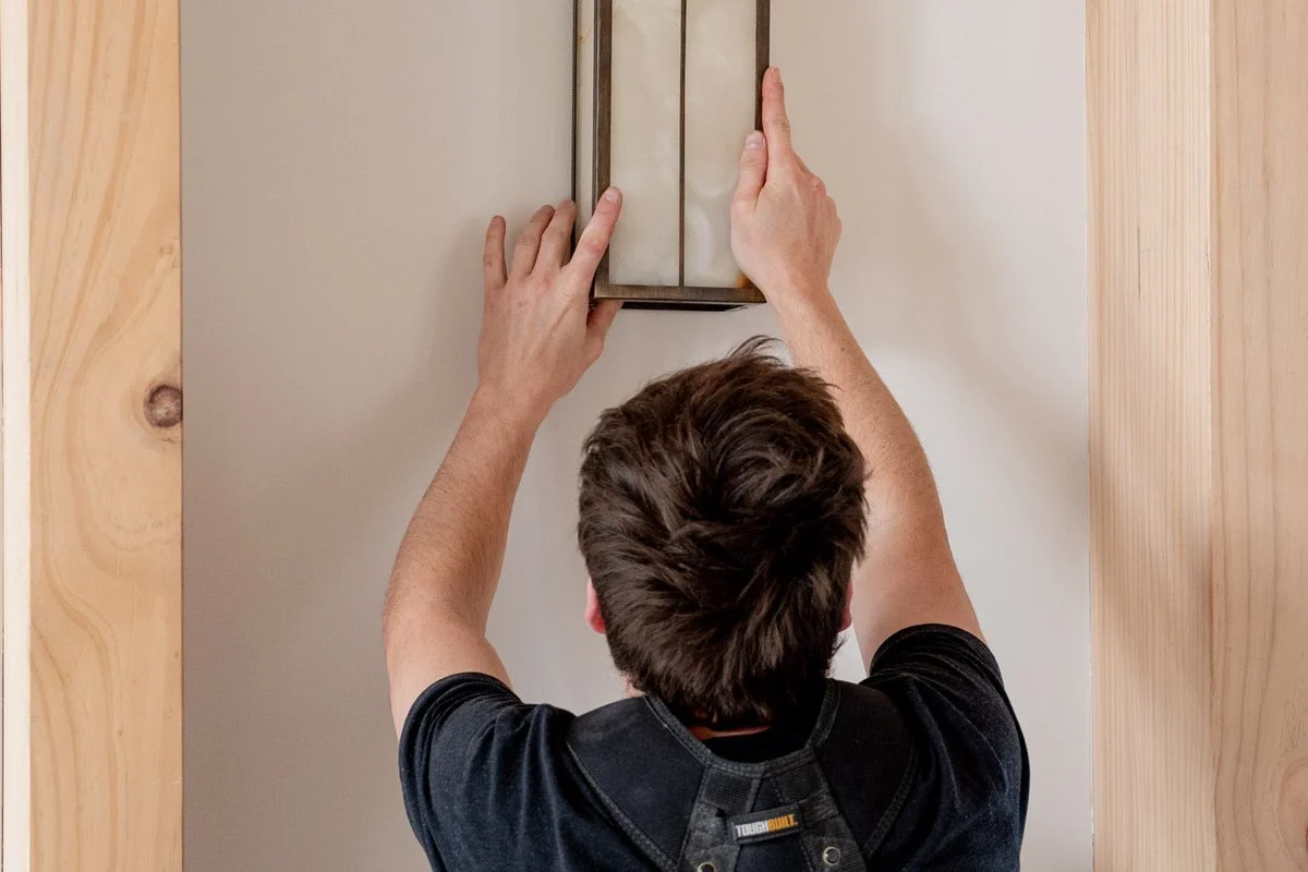 Holding a framed wall light up against a wall between timber posts, back turned.