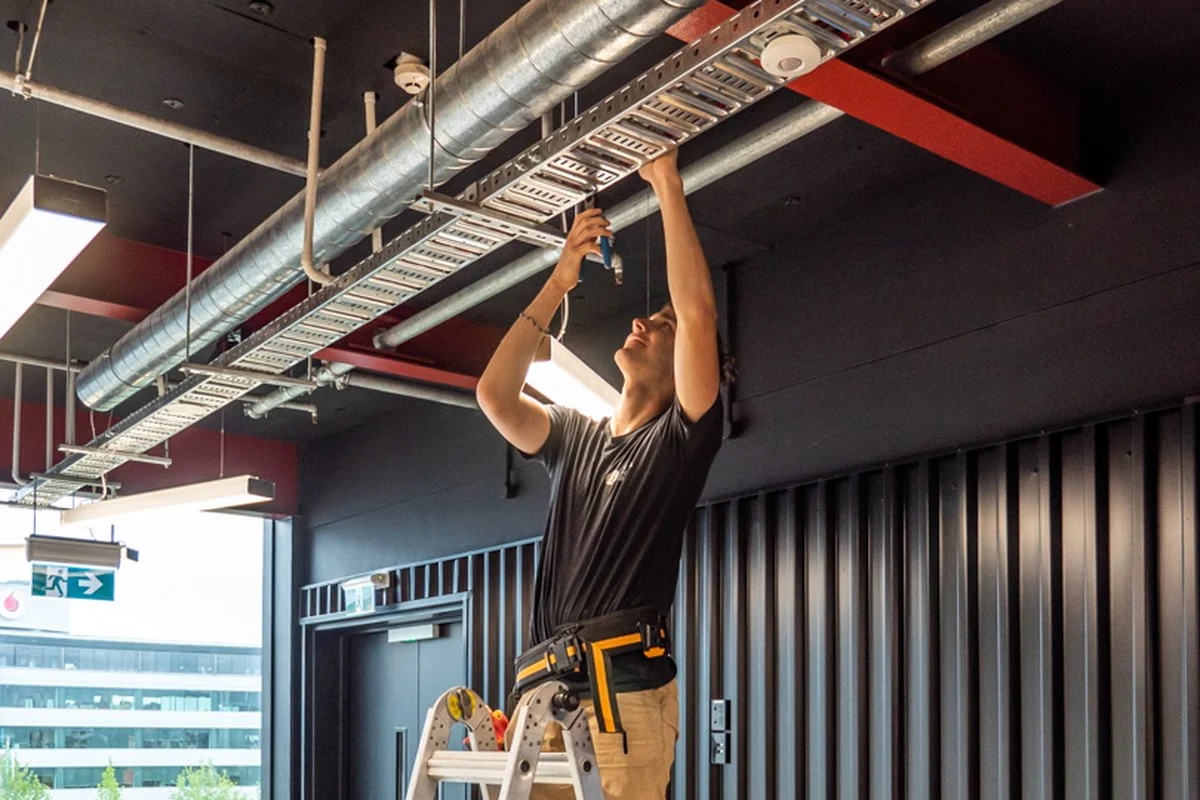 A worker on a ladder reaching up into cable tray and ductwork.