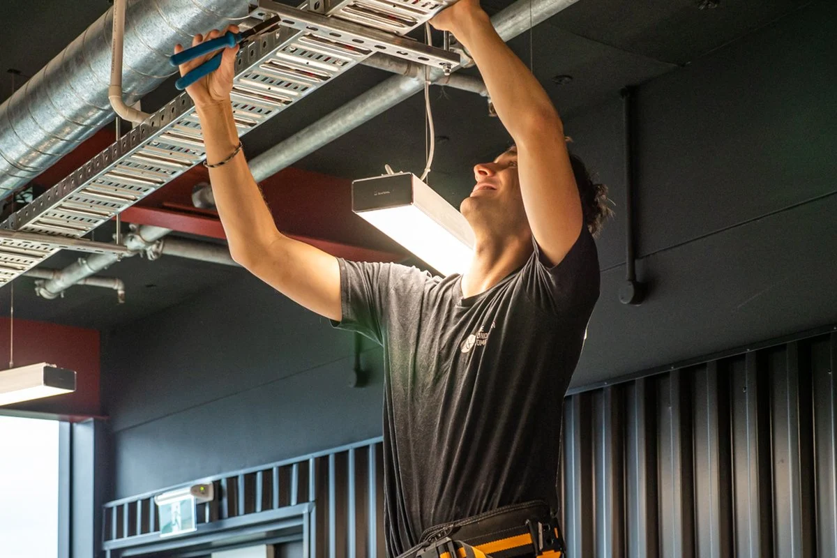 A worker on a ladder reaching up into a cable tray with a hand tool.