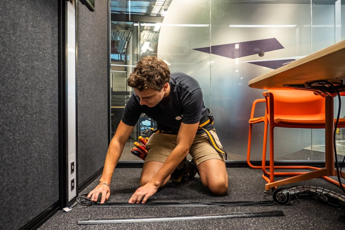 Kneeling on a floor, running cable into a duct beside a glass partition.