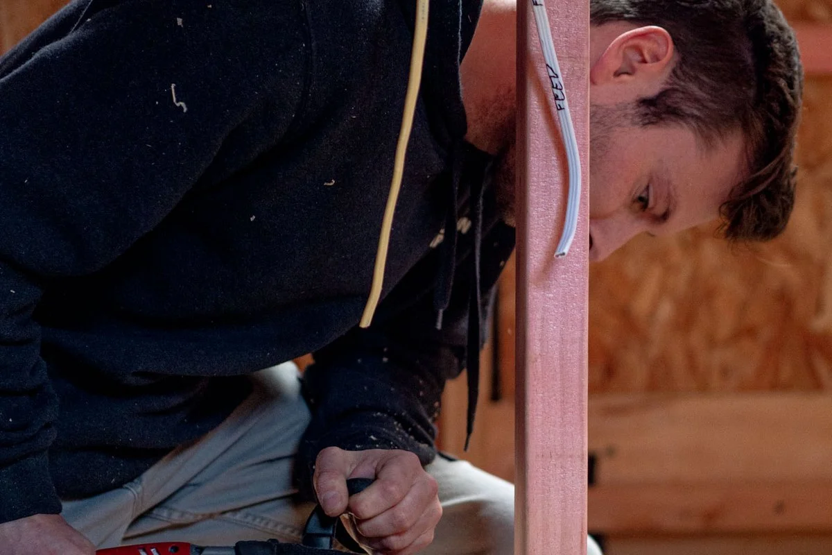Kneeling on a plywood floor, drilling a bottom plate, cables hanging alongside.