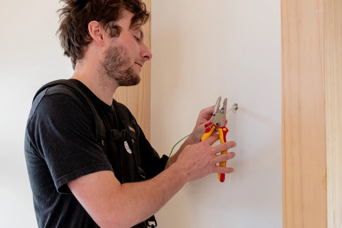Using pliers on a cable at a wall point, tool belt loaded, head down.