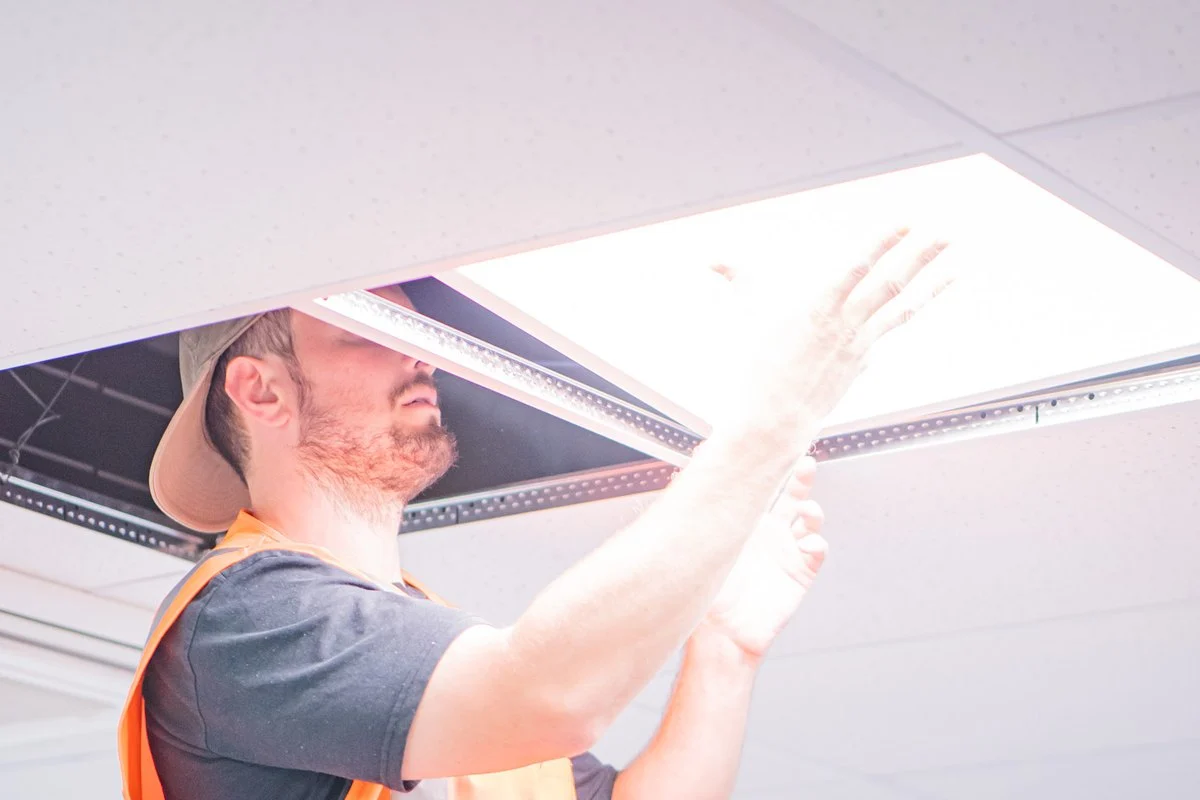 Lifting a lit LED panel into a suspended ceiling grid, both arms raised.