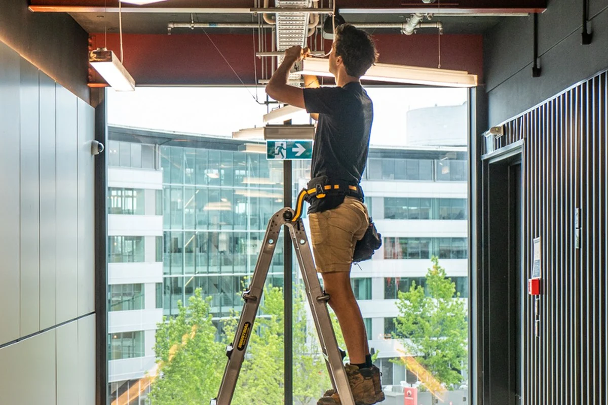 Standing high on a ladder fitting a linear LED batten in a lobby ceiling.