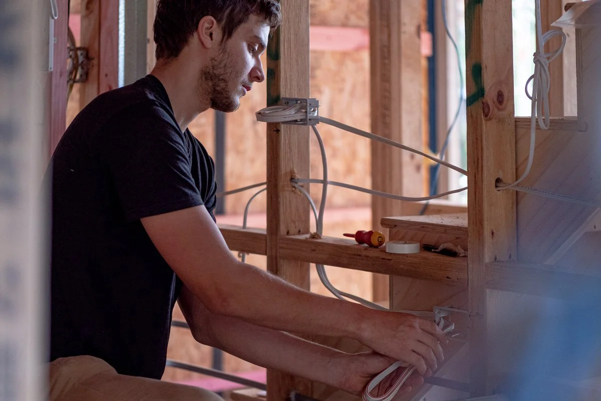 Kneeling at a stud wall, both hands on an outlet box, timber framing around.
