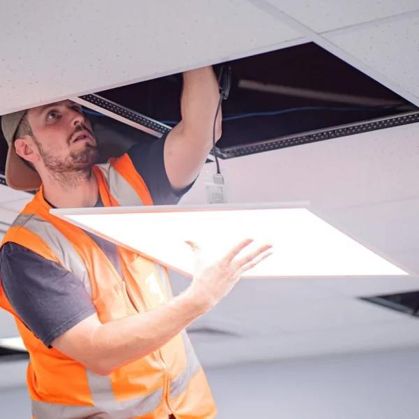 An electrician in a high-vis vest fitting an LED panel light into a suspended ceiling grid