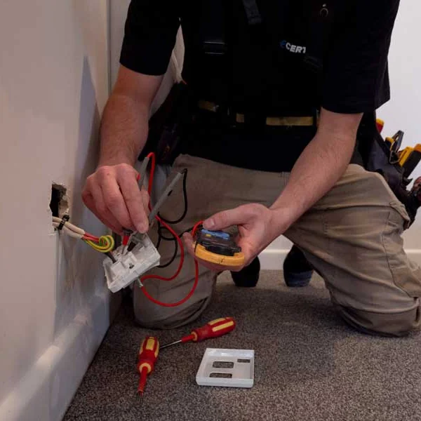 An electrician kneeling at a skirting board, testing a socket with a multimeter